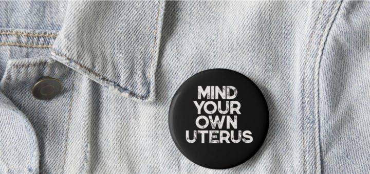 A closeup of a black pin that says "MIND YOUR OWN UTERUS" on a pale blue denim jacket