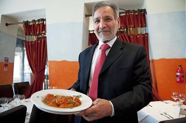 Ali Ahmed Aslam, with white hair and a beard, wearing a dark suit and a red tie and holding a plate of chicken tikka masala. 