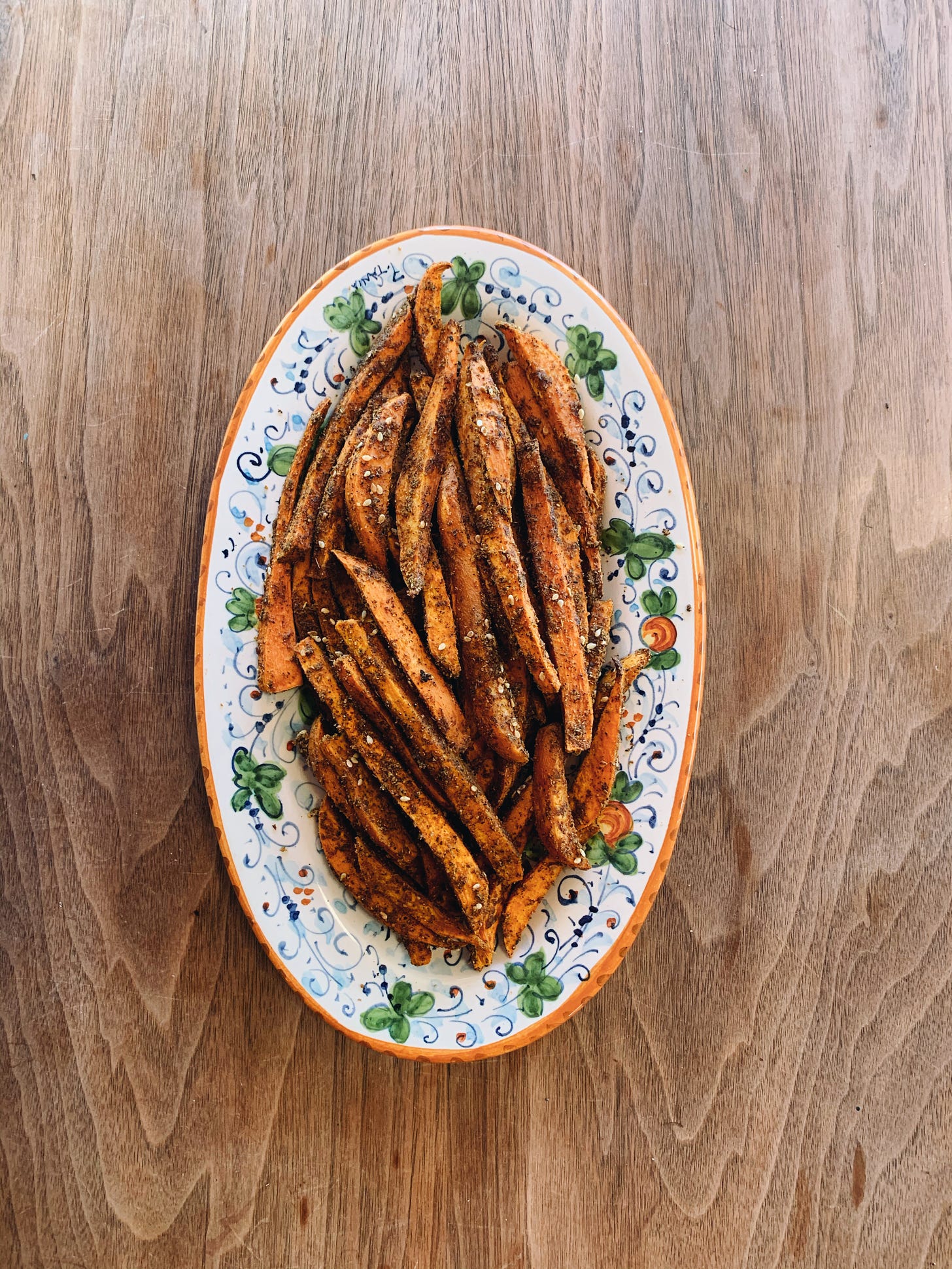 sweet potato fries baked and covered in spices. they are arranged on a decorative plate with an orange rim and green flower designs. the photo is taken from above and the plate is sitting on a table top. 