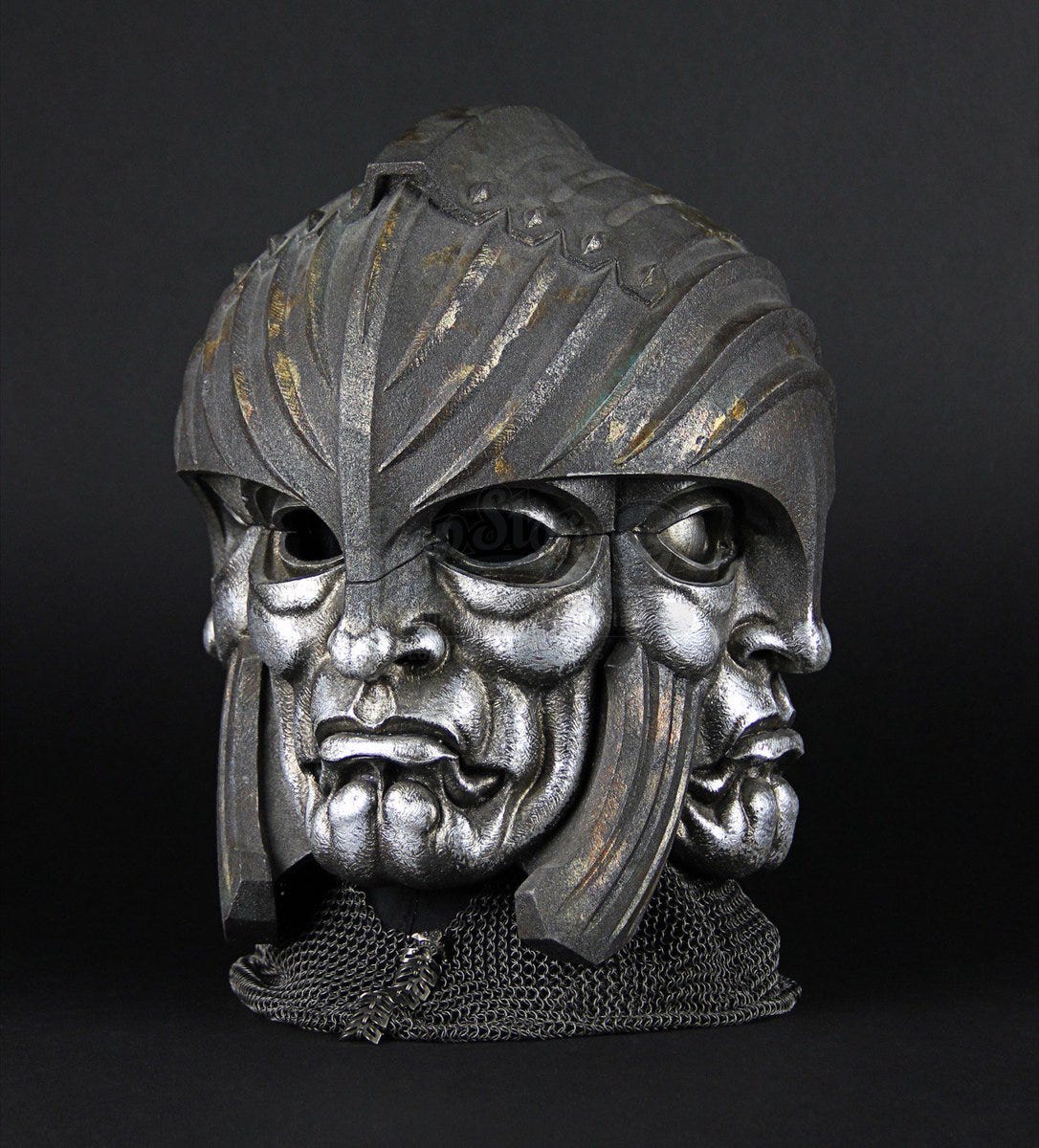 Image Description: A helmet made of glossy silver metal. The front is a face with only eye holes. Each other side of the helmet as the exact same face except no eye holes. The art style is reminiscent of Roman metallurgy and iconography.
