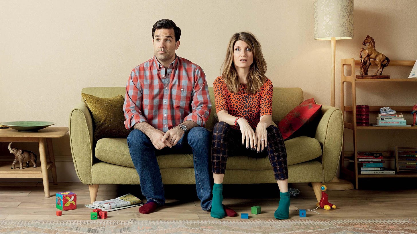 Two people sit on a loveseat with frazzled looks on their face, childrens toys around them on the floor