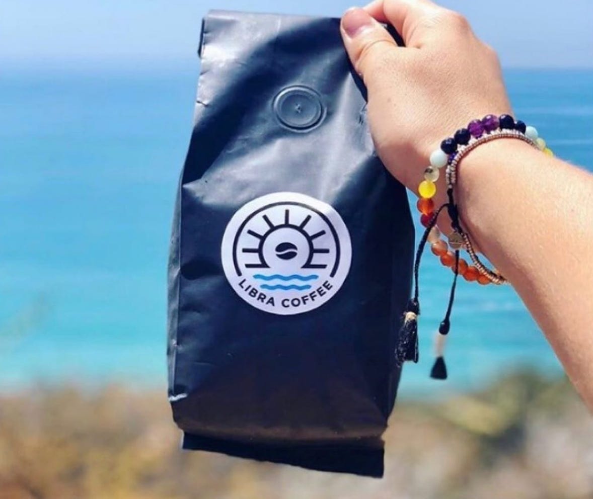 Blue bag of Libra Coffee Beans held up in front of the ocean. Water is bright turquoise. Coffee bag is navy. 