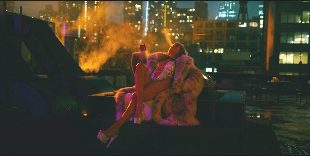 JLo lying resplendent on a roof in the film Hustlers. She's smoking a cigarette and blowing smoke into the air while wearing a silver leotard and enormous fur.