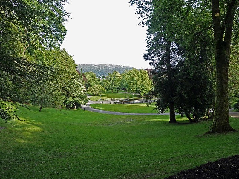 Image description: A green grass slope with trees on both sides allow for a visita of the park walkways below.  