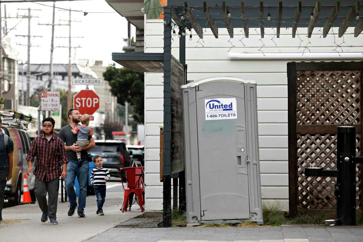 People are seen walking on 24th Street next to a portable toilet at Noe Valley Town Square on Tuesday, October 18, 2022 in San Francisco, Calif. Noe Valley Town Square will be the site of the construction of a toilet stall that will cost $1.7 million and take more than two years to build.