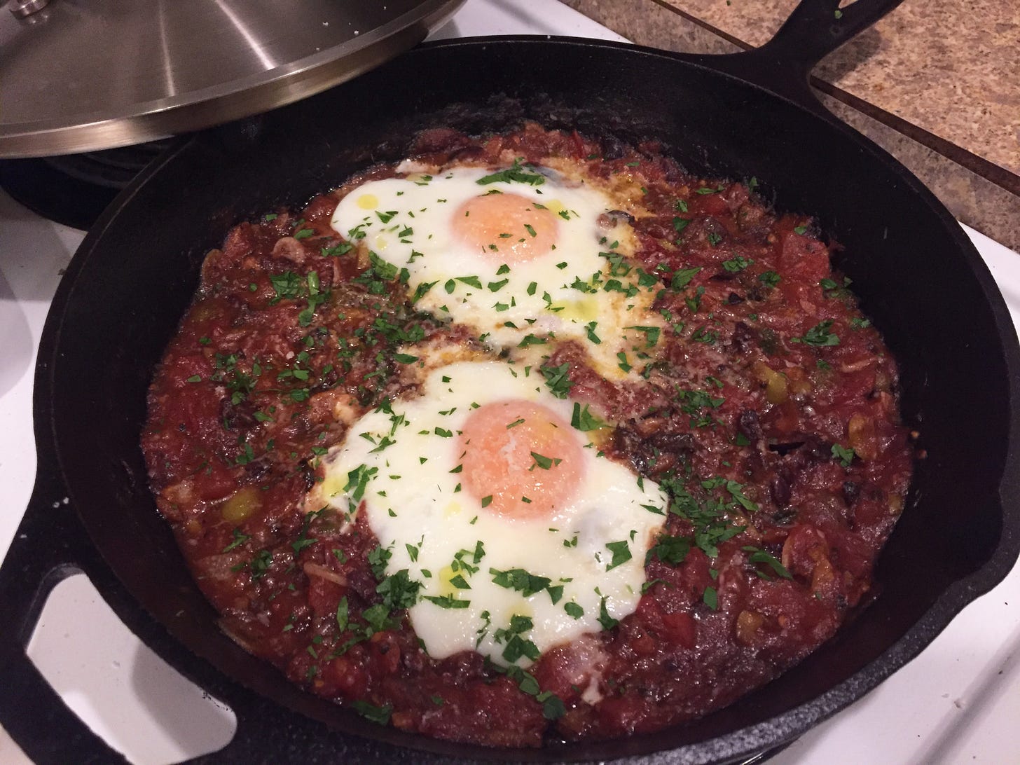 a cast iron pan filled with deep red puttanesca sauce. Two poached eggs sit on top, sprinkled with parsley and parmesan and drops of olive oil.
