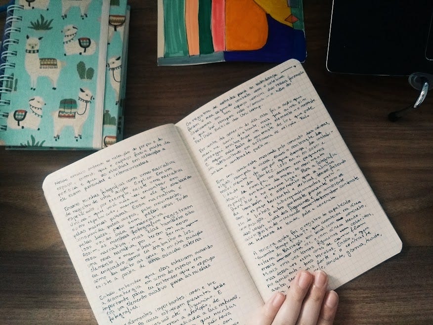 Imagem vista de cima de uma mesa de madeira com um caderno aberto em duas páginas escritas a caneta preta. O caderno está sendo segurado por uma mão branca. Ao redor do caderno, tem um outro caderno colorido, um desenho e um computador.