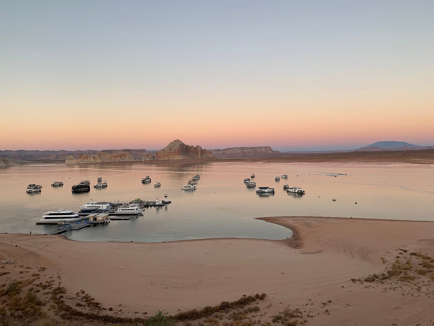 The sun sets in hazy orange and pink over a bay where houseboats dot the horizon and desert rocks rise in the background