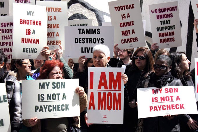 Several dozen middle aged moms stand outside with protest posters reading I AM A MOM, and My Son is Not a Paycheck.