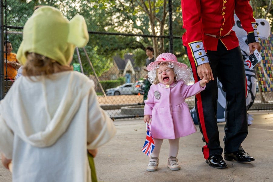 A young child in a Queen Elizabeth Halloween costume cries while holding the hand of an adult dressed as a palace guard.