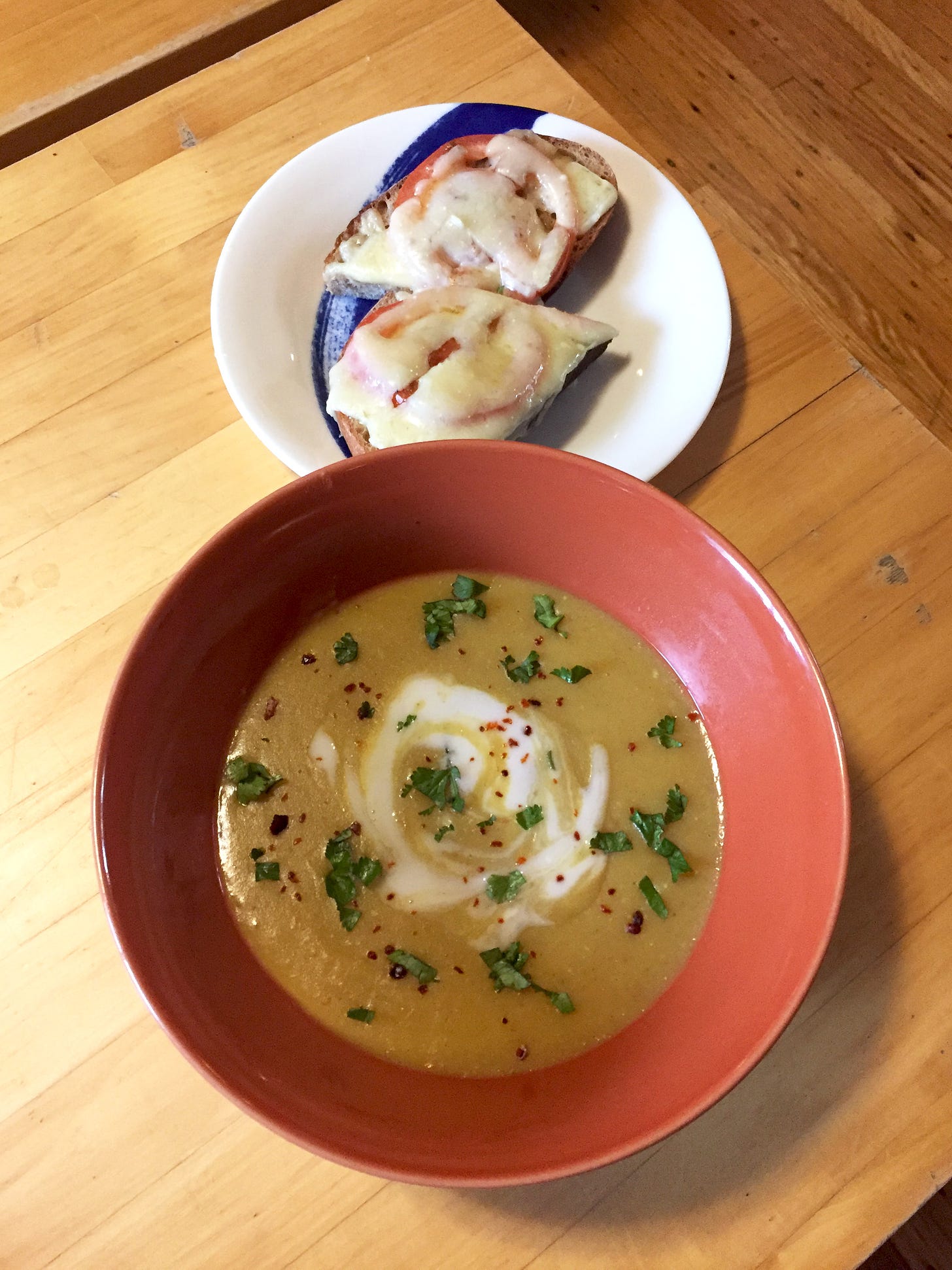 In an orange bowl, a thick and deep yellow soup swirled with coconut yogurt and sprinkled with cilantro and chili flakes. On a plate behind it and to the right is a slice of cheese toast with tomato, sliced in half.
