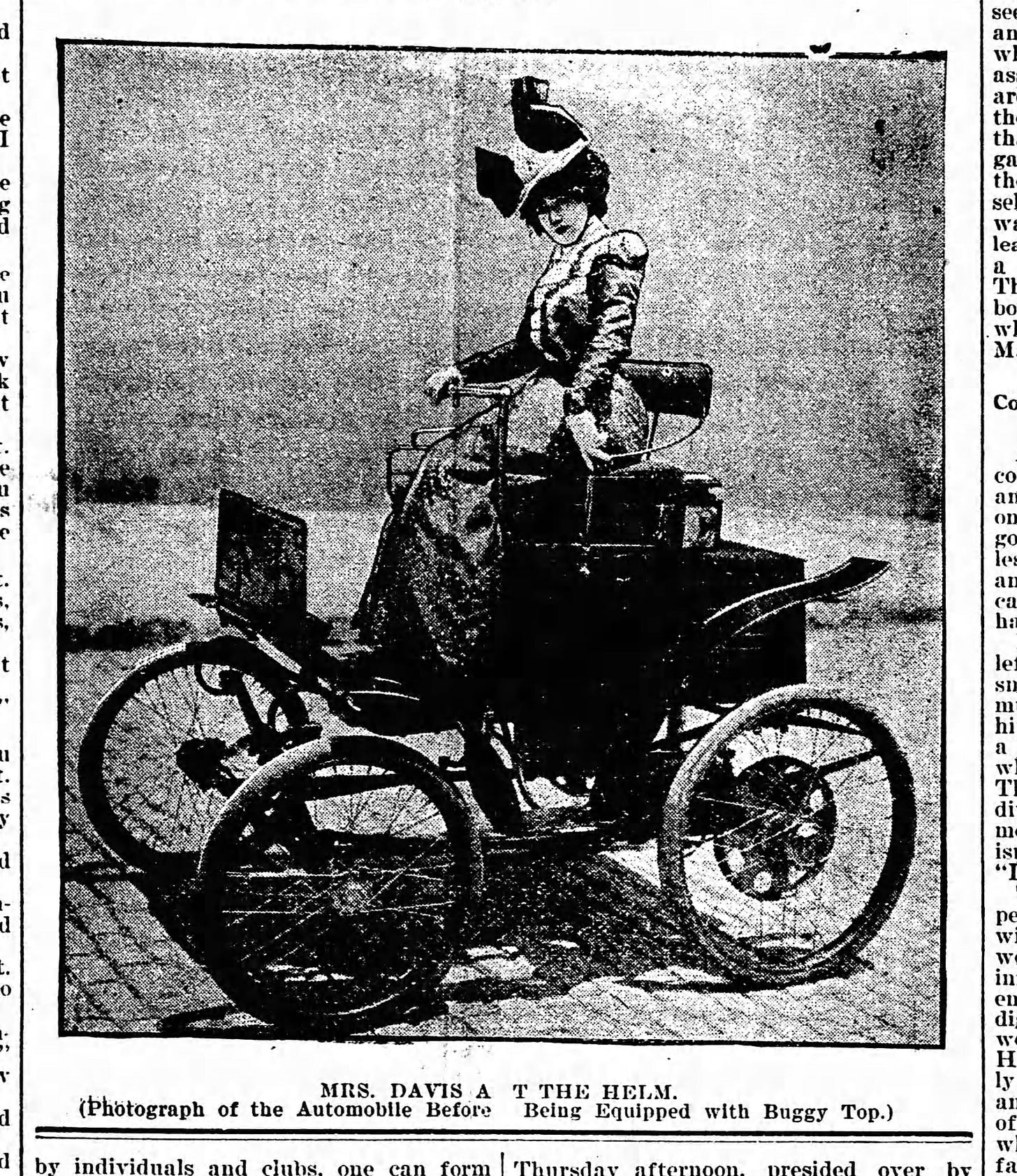 Photo from The Virginia Enterprise (Virginia, Minnesota, Friday, July 21, 1899) of a woman in period clothing (a dress covering the ankles, tight waist, closed collar, long sleeves, and fashionable hat) sitting at the helm of a Duryea automobile. The Duryea resembles horse-drawn carriages of the era and features four spoked wheels that look like wheels from a bicycle, a stylish padded seat, and a tiller for steering. Caption reads, "MRS. DAVIS AT THE HELM. (Photograph of the Automobile Before Being Equipped with Buggy Top.)"