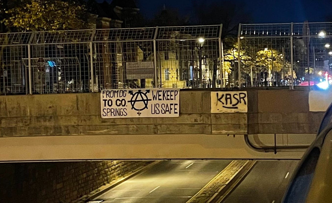 Banner hanging from overpass that says "From DC to Co. Springs we keep us safe", as well as a trans symbol superimposed on a circle A