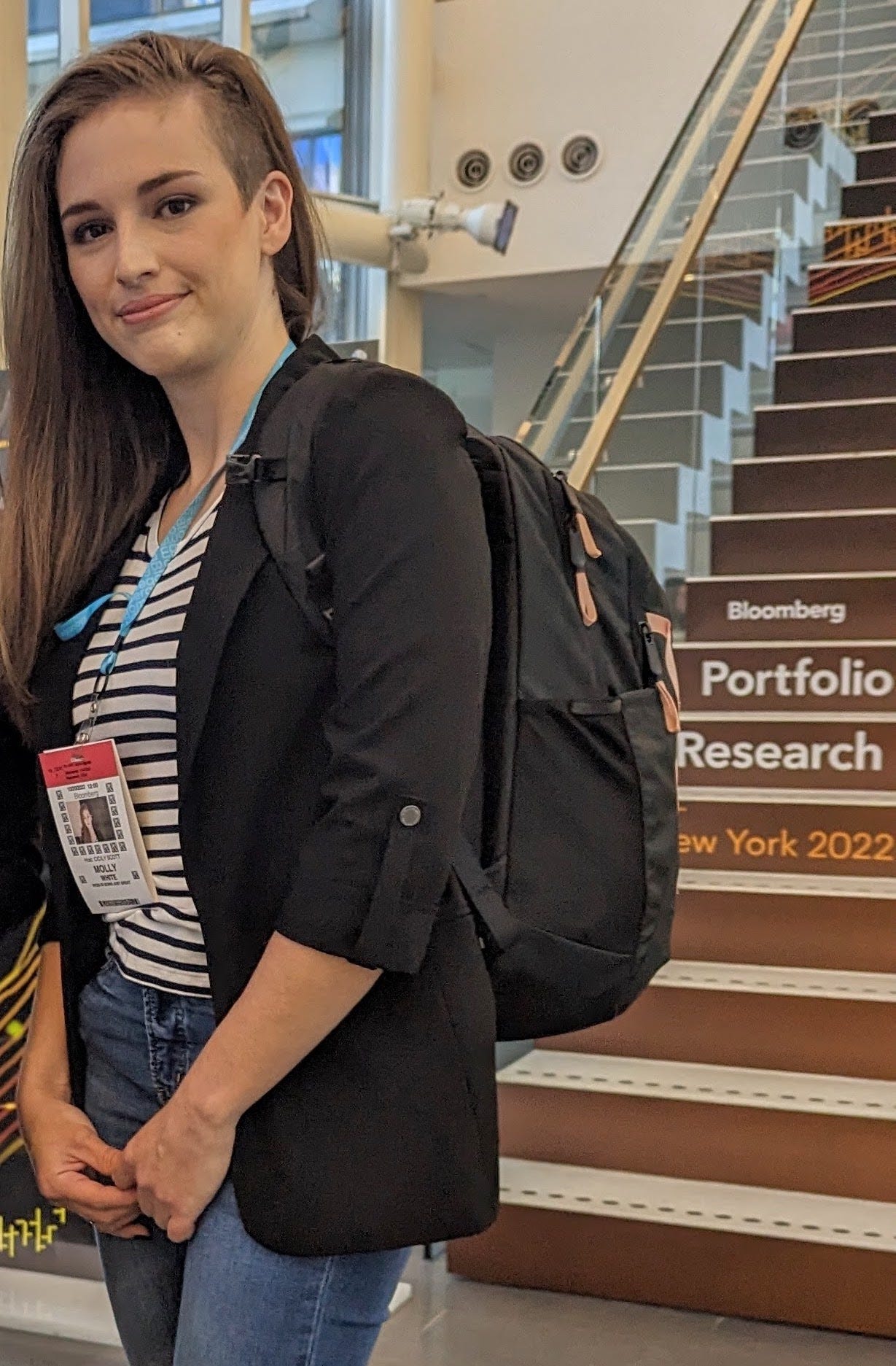 Photo of Molly standing in an office building. She's wearing a white and navy striped shirt, a black blazer, and jeans. Behind her is a large staircase with the text "Bloomberg Portfolio Research New York 2022" printed on the risers. Photo of Molly standing in an office building. She's wearing a white and navy striped shirt, a black blazer, and jeans. Behind her is a large staircase with the text "Bloomberg Portfolio Research New York 2022" printed on the risers.
