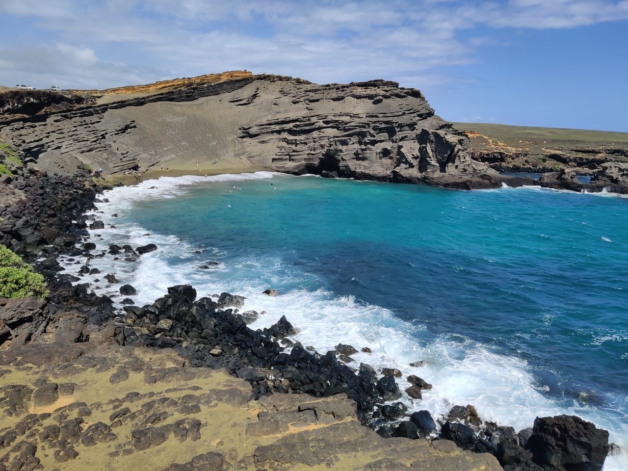 A horseshoe shaped beach with olive sand and brown cliffs and rocks, with a vibrant blue waters crashing against them.