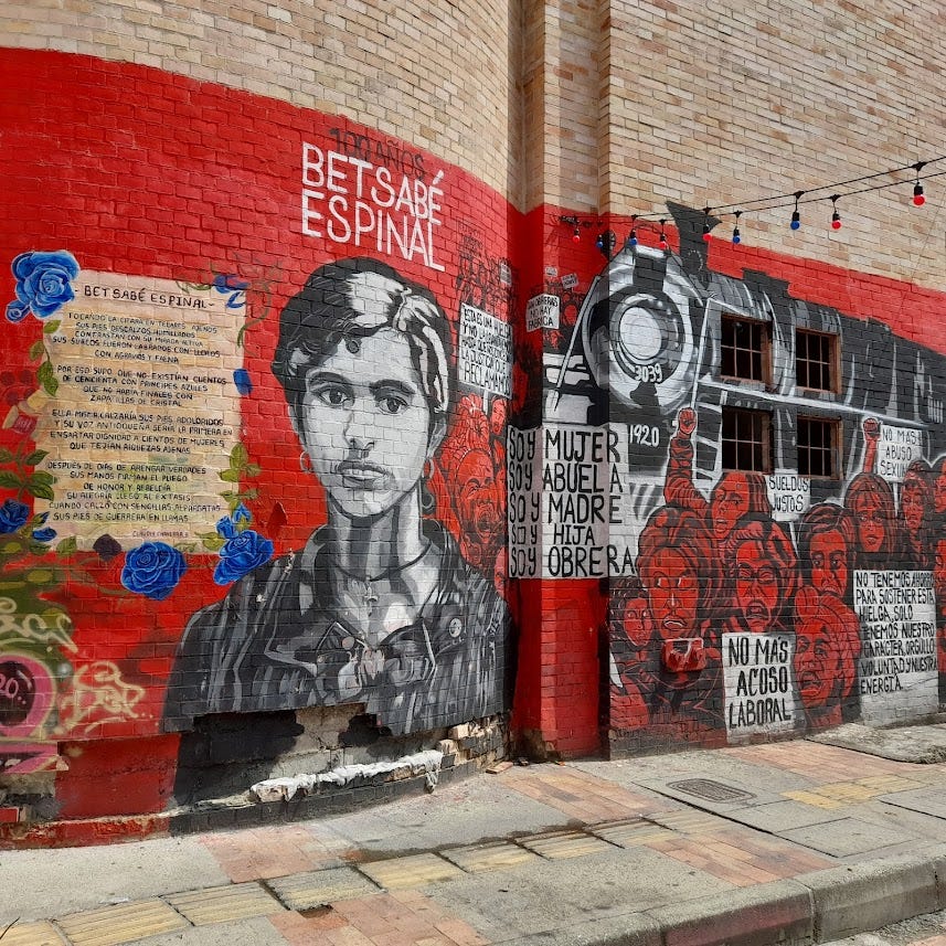 Graffiti memorializing Betsabé Espinal in her birthplace of Bello. A red wall, overlaid with a poem in her honour, her image in black and white, a train, and protesters with signs representing the causes she fought for.