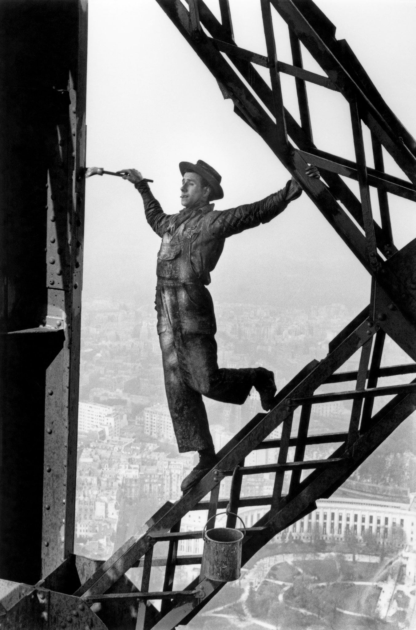 a man gingerly hangs onto a girder on the Eiffel tower, high above the ground, a cigarette in his mouth and a paint brush in his other hand