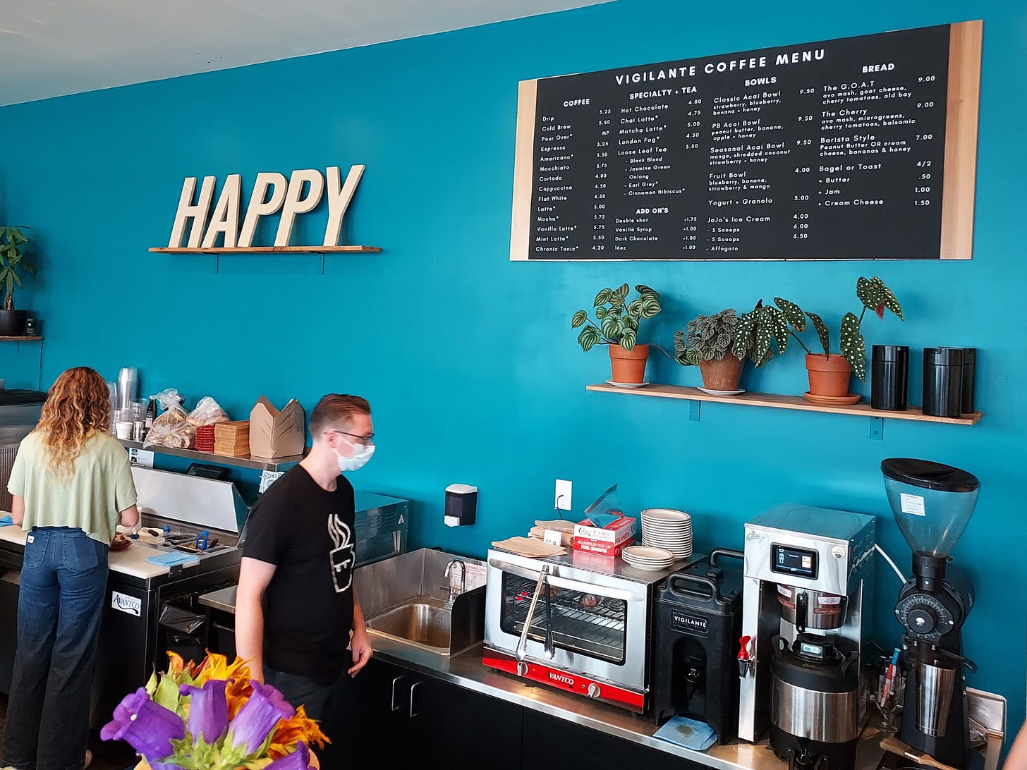 The word "HAPPY" in white, wood, block letters is propped up against a teal blue wall on a shelf next to a black coffee shop menu over the back bar of a cafe. Employees prep food with their back to the camera.