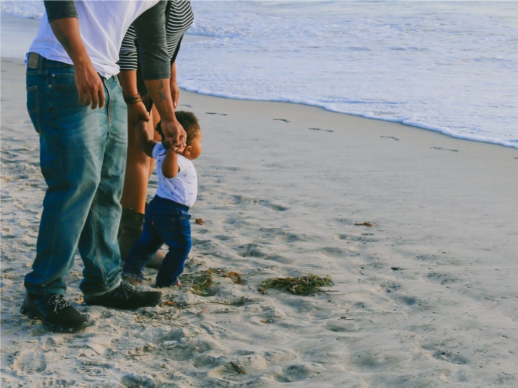 Photo of a couple on the beach holding their toddler’s hands as they look out to sea, by Larry Crayton on Unsplash