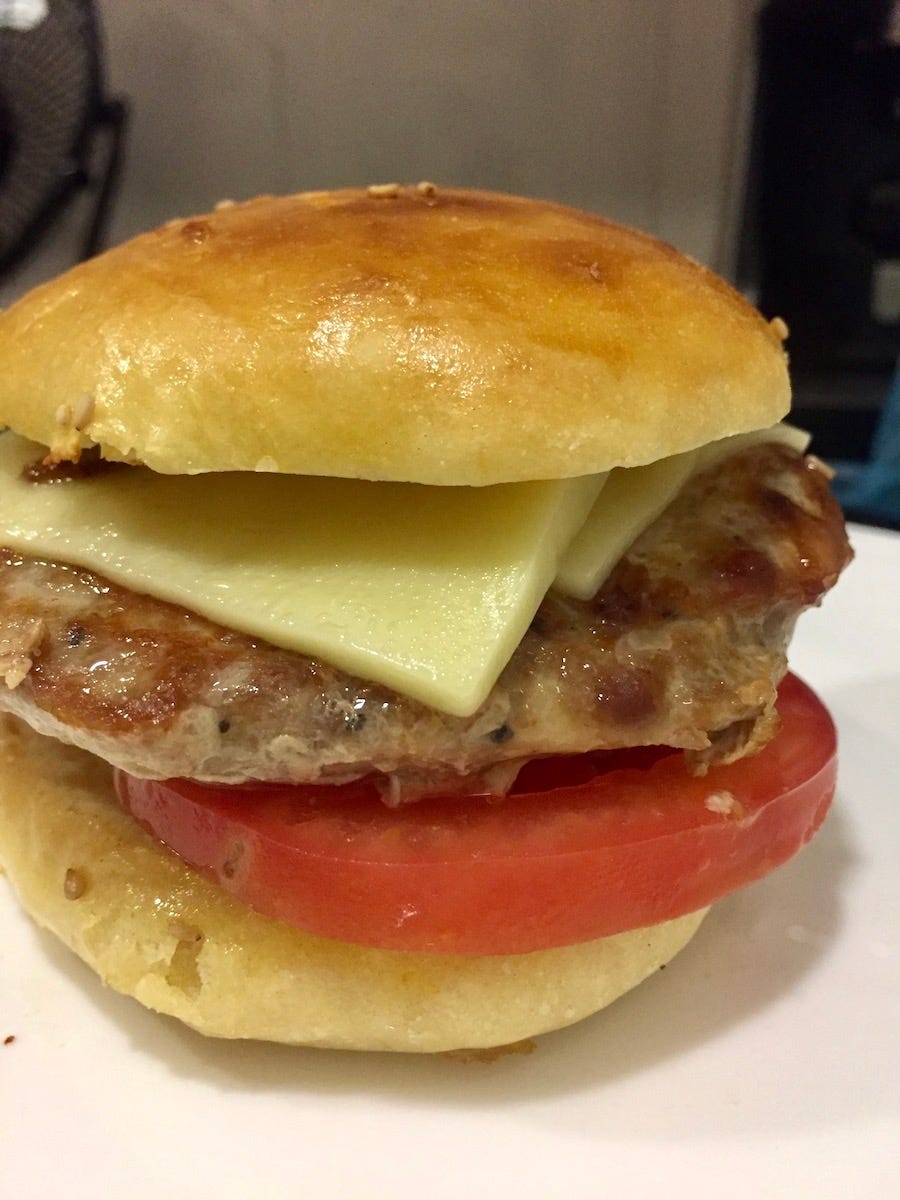 photo of close up of a pork burger with homemade burger bun, two slices of cheddar cheese, a thick slice of tomatoe