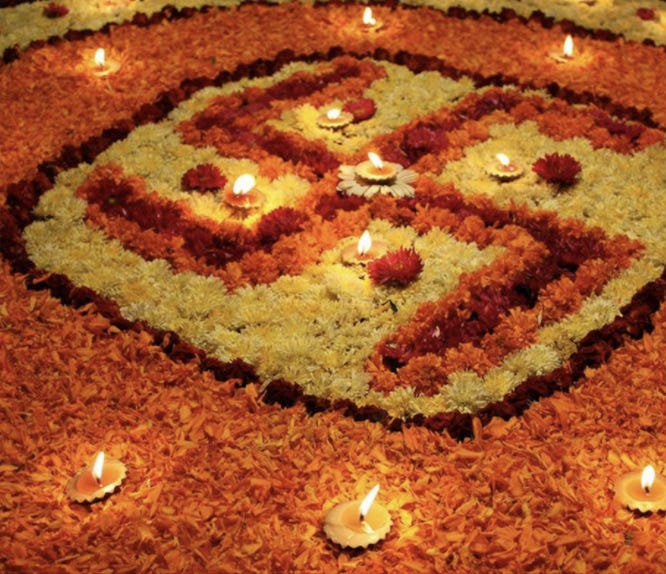 A swastika symbol designed with orange, red, and yellow flowers and lamps. The red flowers are the right angles of the symbol, bordered with orange flowers, and surrounded by yellow flowers. The four dots are represented by four red flowers and four lamps. The whole symbol is surrounded by a red flower border, which is itself surrounded by orange flowers and more lamps, with another red flower border. That is surrounded with more yellow flowers and lamps.