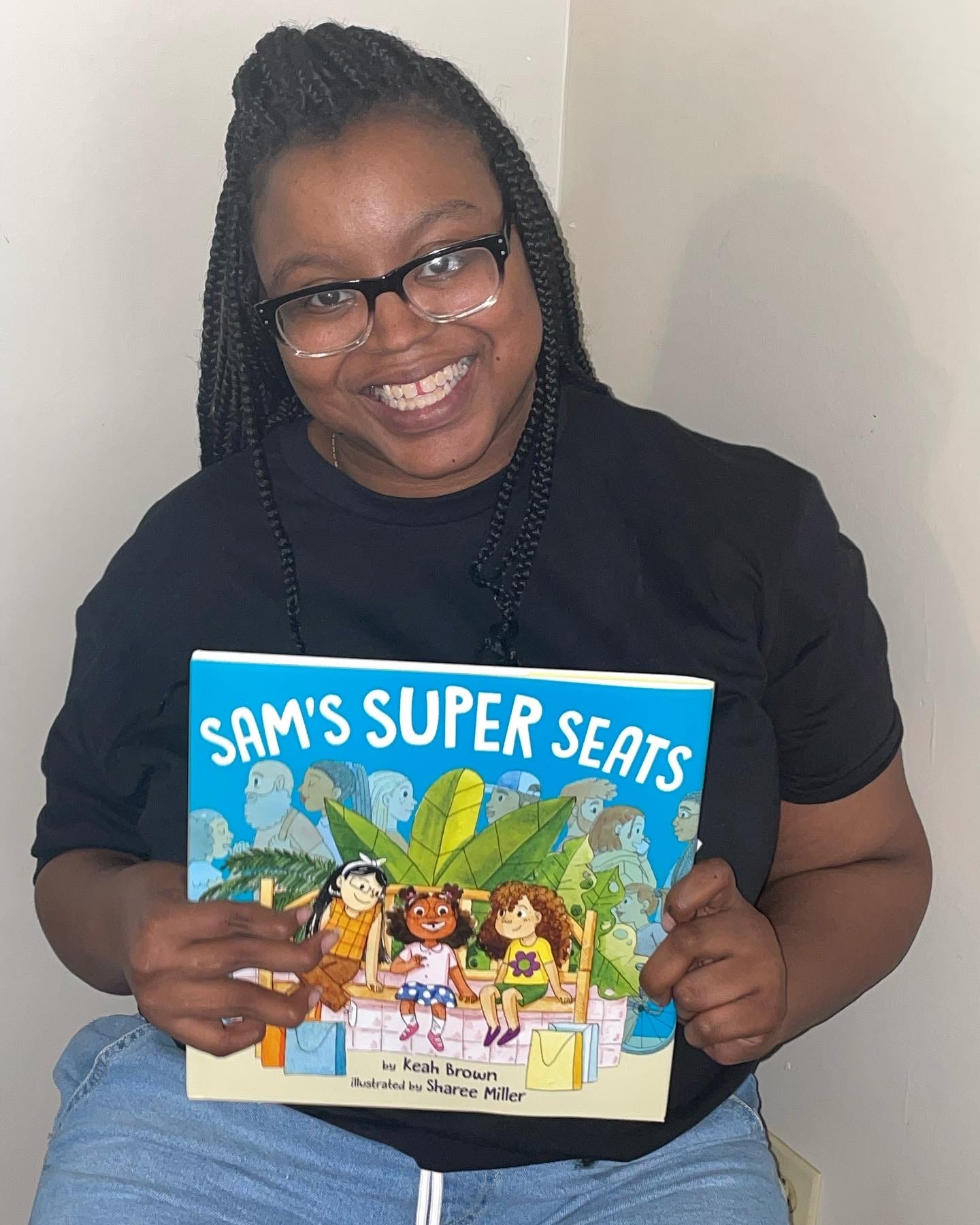 Keah smiles at the camera wearing black and white glasses. Her braided hair is  up in a ponytail and she is wearing a black shirt and blue Jean shorts. She is holding the physical copy of Sam’s Super Seats in her hands.