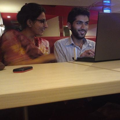 Sidra and Waqas sitting next to each other in a café. They are looking at a laptop, they are both smiling and overall seem to have a good chemistry.