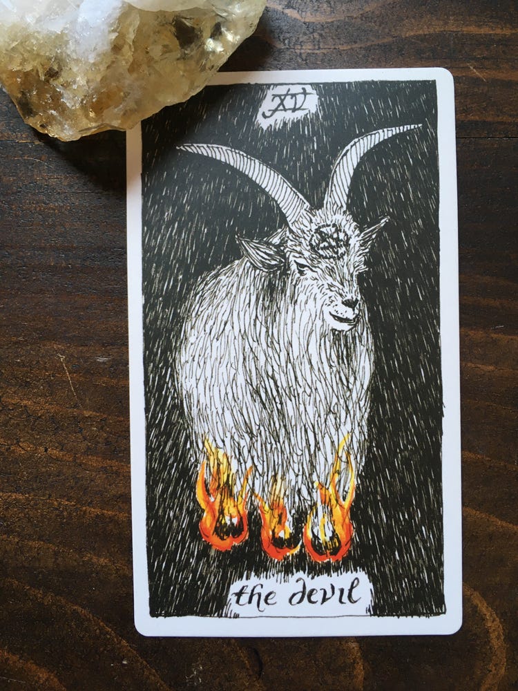 A tarot card held down by a citrine crystal. There is a line drawing of a goat-like creature with huge horns, an upside-down pentacle on their forehead, and hooves on fire. The card is all black and white except for the flames. The number 15 is at the top, in roman numerals, and at the bottom, the text is “the devil.” 