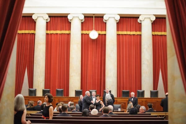 People in formal attire sit on and stand amid the audience benches that face the bench where the justices sit in the Supreme Court chamber. Behind the justices’ bench are red curtains and four white marble columns.