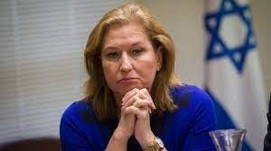 Tzipi Livni - wearing a blue top, hands clasped, staring thoughfully, with the israeli flag behind her