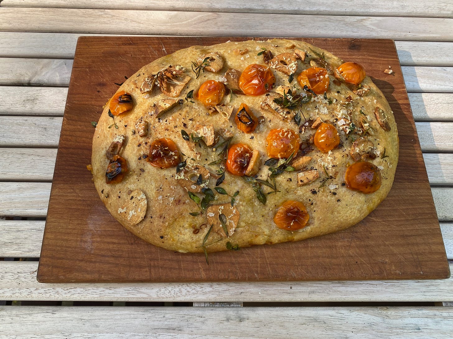 An oval focaccia loaf on a wooden cutting board. It’s topped with blistered cherry tomatoes, thinly sliced fried garlic, herb springs, and grated Parmesan.