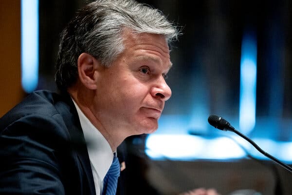 Christopher A. Wray, the F.B.I. director, wearing a blue suit and facing a microphone. 
