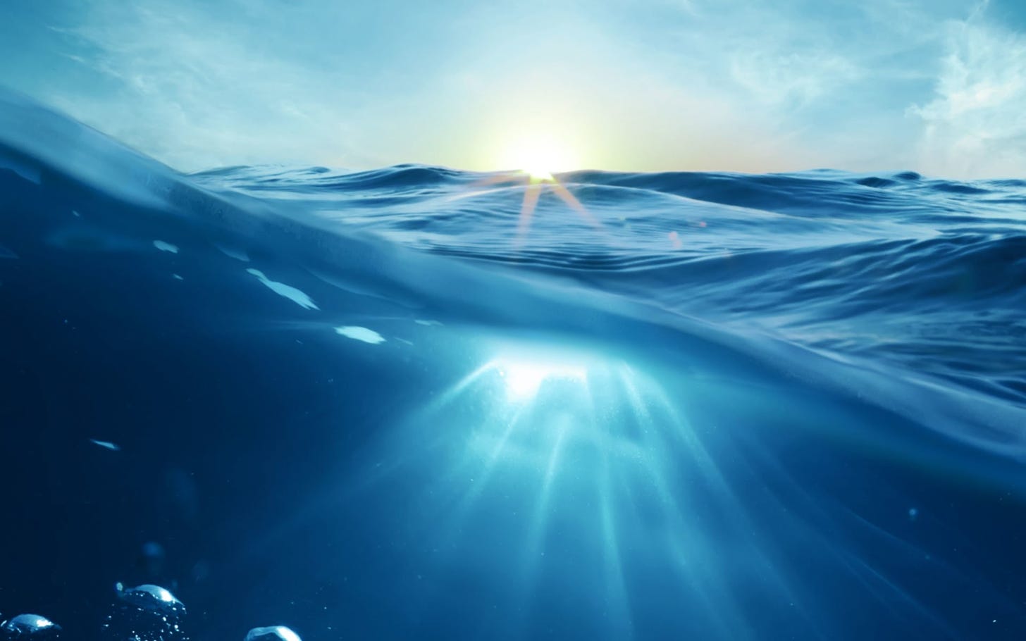 Imagem é uma fotografia capturada dentro da água, com parte da lente submersa registrando o movimento da água, bolhas e raios de luz penetrando nela, e outra parte registrando marolas com o céu e o sol ao fundo.