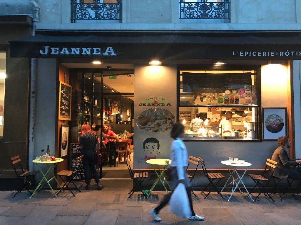 A woman carrying a white plastic bag walks by Paris restaurant JeanneA at night. The restaurant has sidewalk tables.