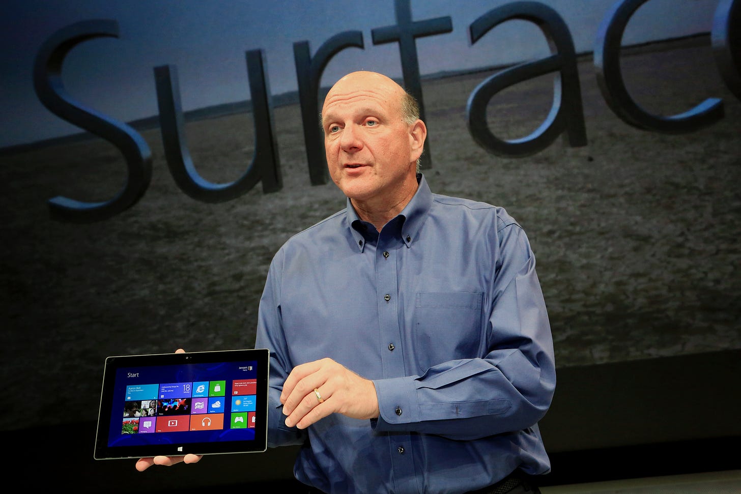 Ballmer is holding a surface on stage. The backdrop is projecting Surface over a field of green grass.