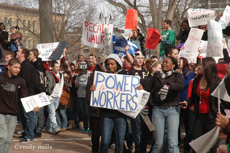A group of Black, Brown, and white high school students march to the Capitol, holding signs that say "Power to the Workers" and "Recall Walker," etc.