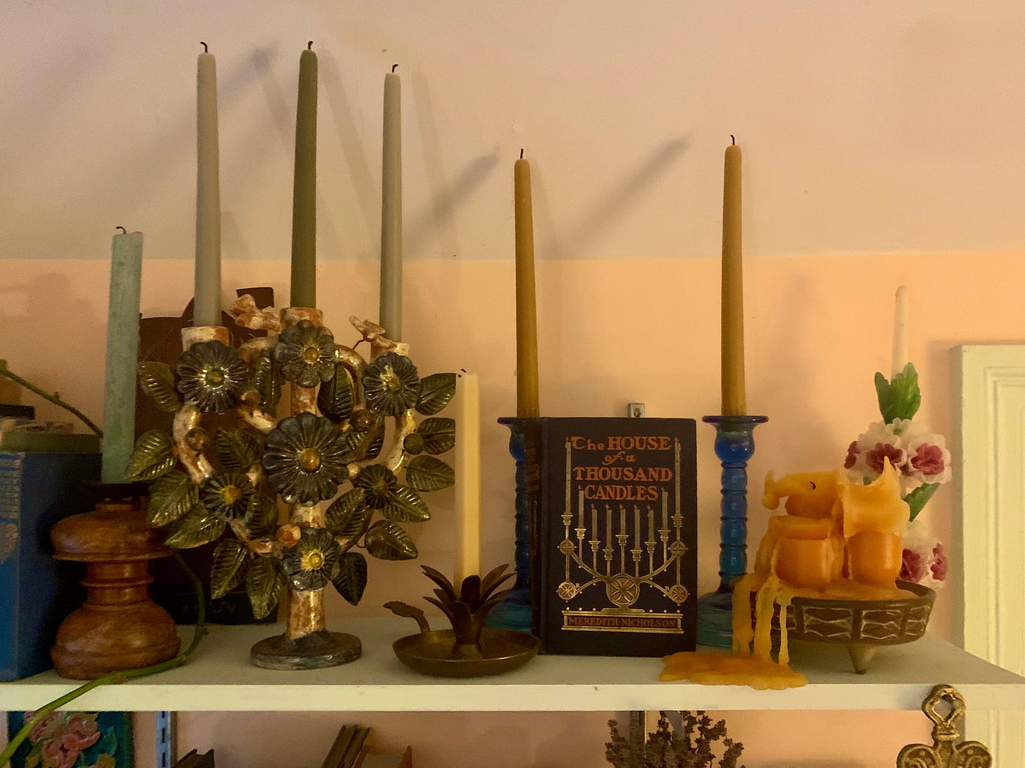A shelf with nine candles in candleholders of many styles and a book titled The House of a Thousand Candles