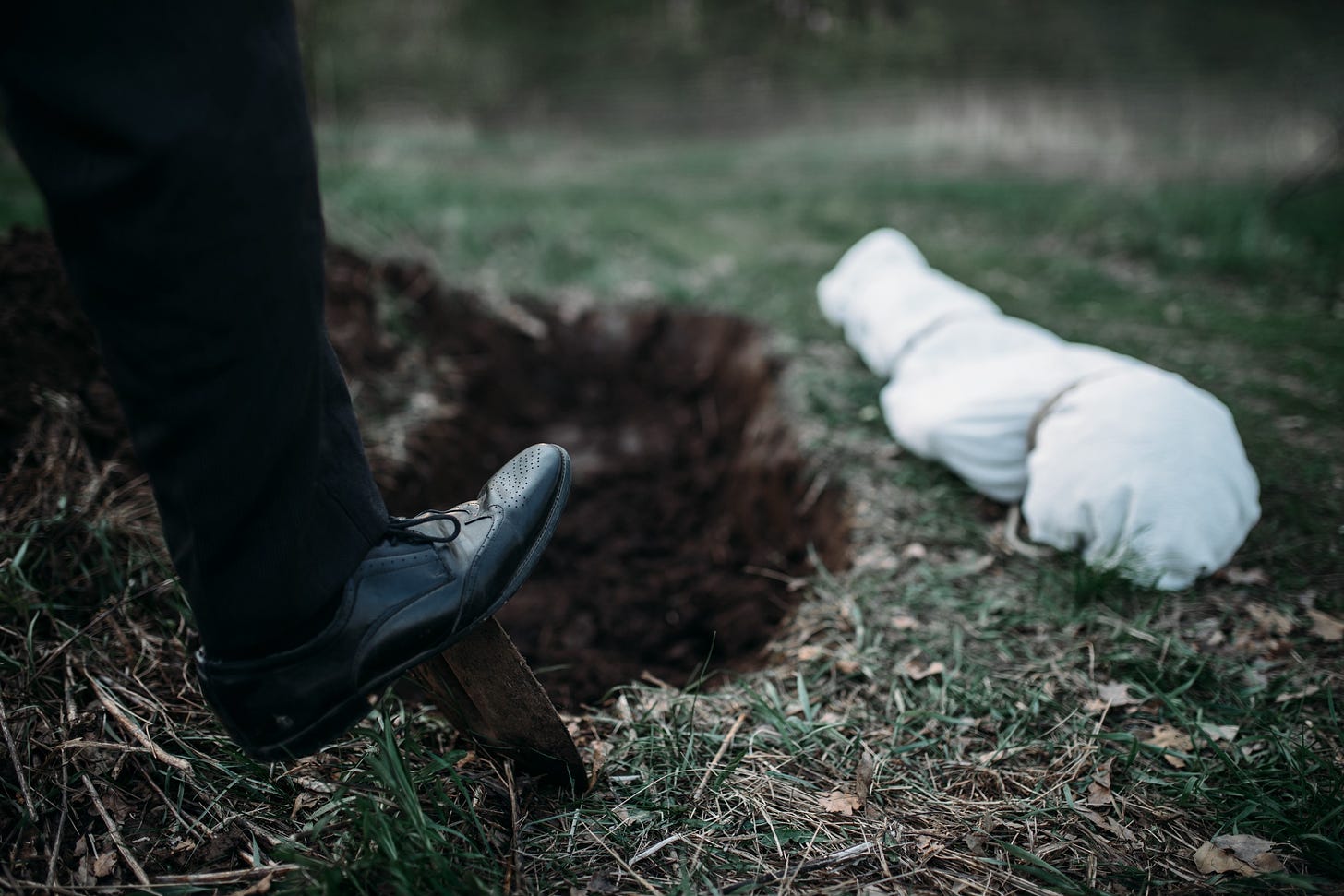 A man in dress shoes and slacks buries a wrapped body in the wilderness.