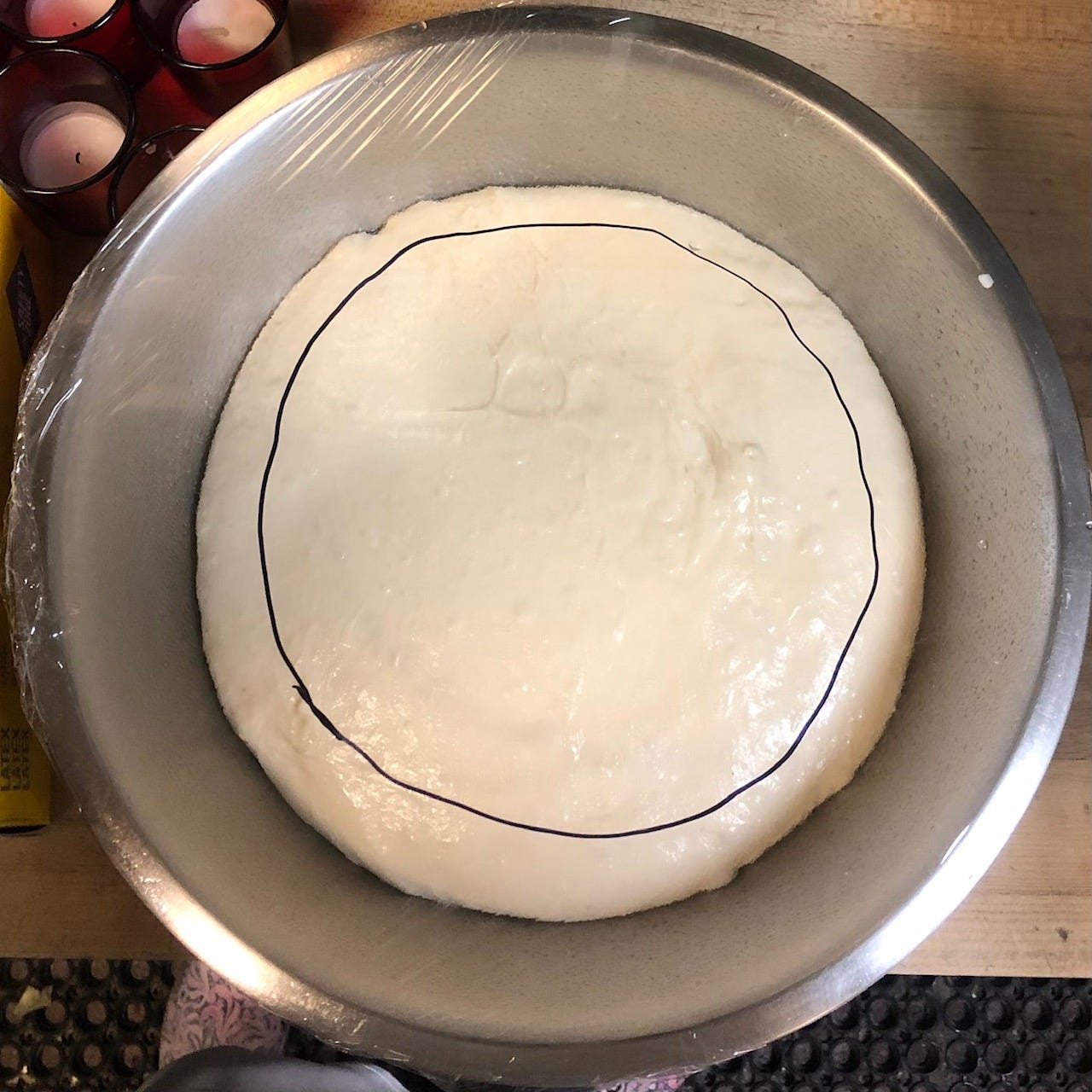 A round of raw dough in a metal bowl, covered with plastic wrap. A black circle drawn on the plastic sits just inside the dough.