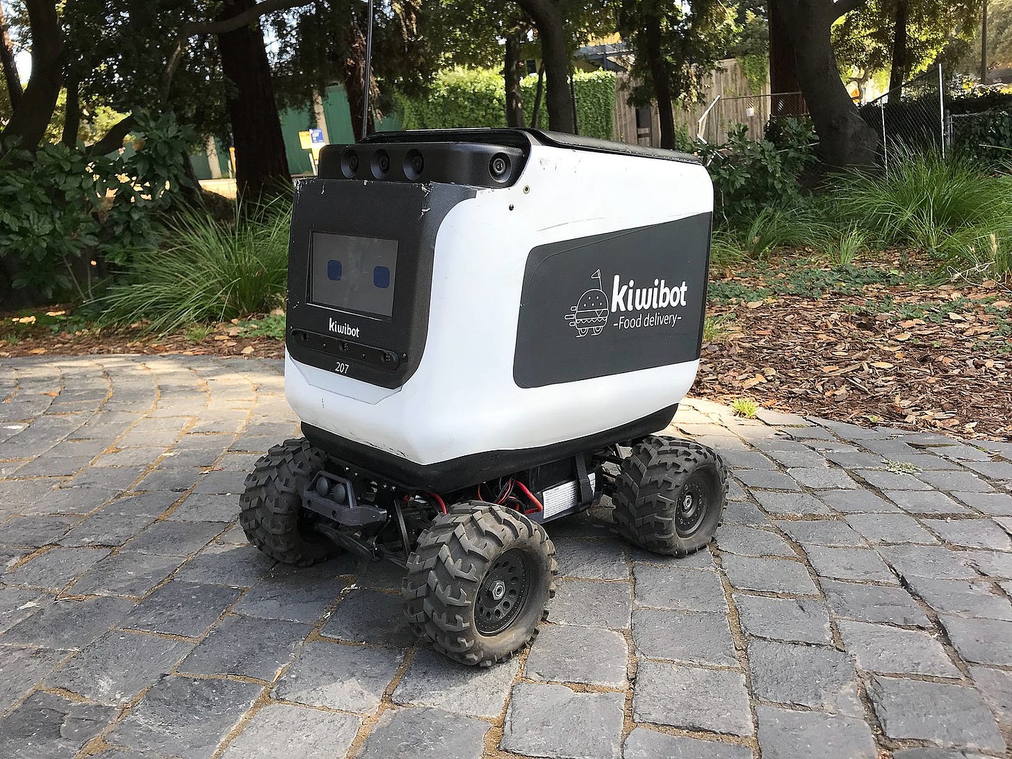 A boxy, four-wheeled Kiwibot food delivery robot seen from a front-facing, three-quarter perspective in a small stone plaza. Its tires have thick, monster-truck-style tread.