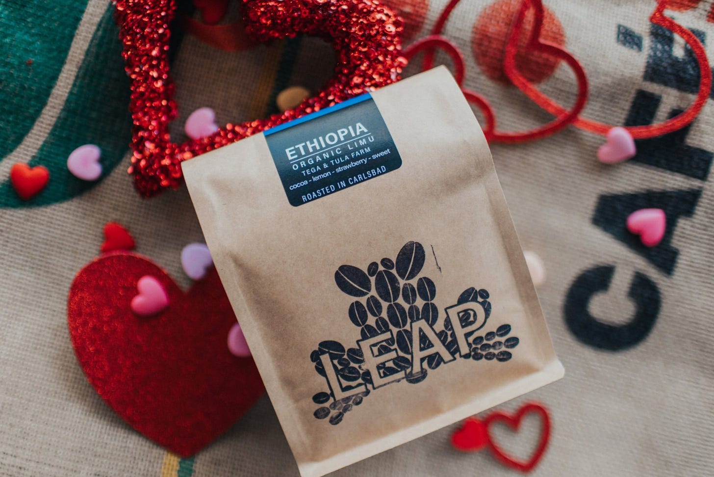 A bag of Leap Coffee Ethiopia beans, a brown bag with the Leap Coffee logo in black ink, resting on a burlap coffee sack surrounded by Valentines day hearts in red and heart candies spread around.