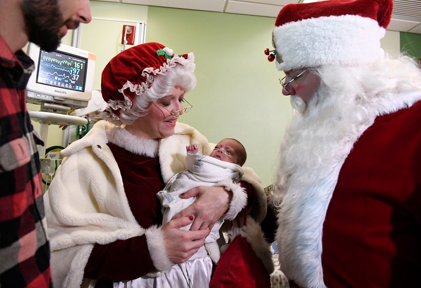 A man dressed as Santa Claus looks on as a woman dressed as Mrs. Claus holds a newborn baby in her arms.