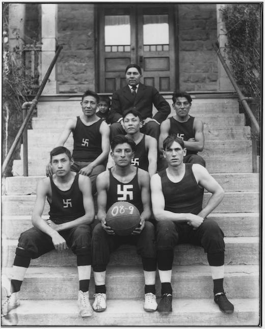 A black and white picture showing young Navajo athletes sitting on steps wearing dark basketball uniform that has a white whirling logs symbol centered on the chest. The athlete at the front and center holds a basketball. And older Navajo man in smart western clothes sits in the center in the back row. Behind them are the doors to a building.