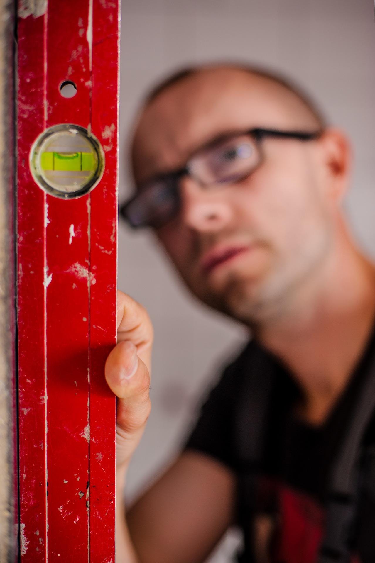 A man is measuring something with a level, which is bright red and clear in the foreground. The man's serious, bespectacled face is blurred in the background.