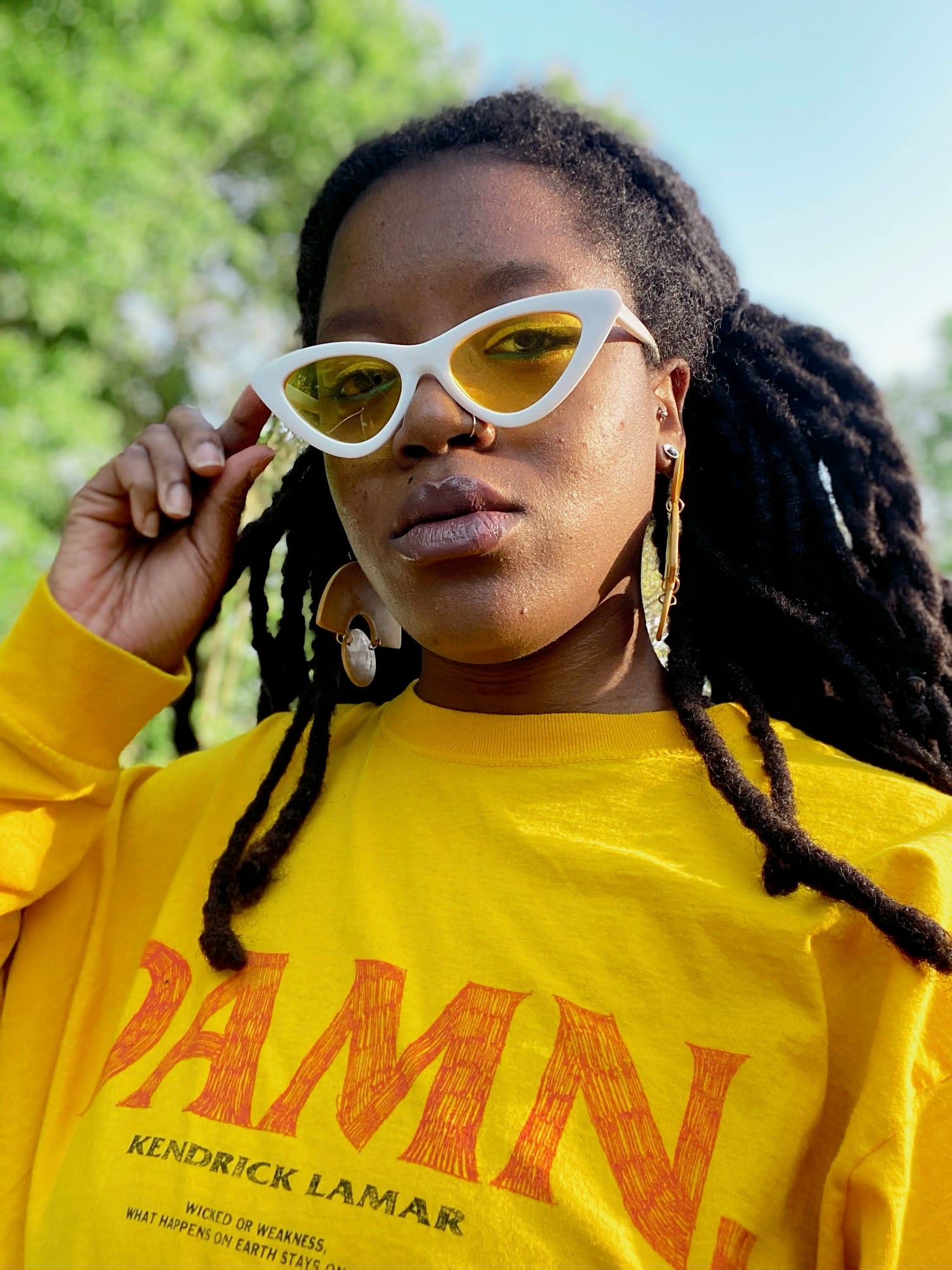 The writer, Tay, wears a yellow t-shirt that says "DAMN." from rapper Kendrick Lamar's 2017 tour. Tay also wears white sunglasses with yellow lenses while staring at the camera.