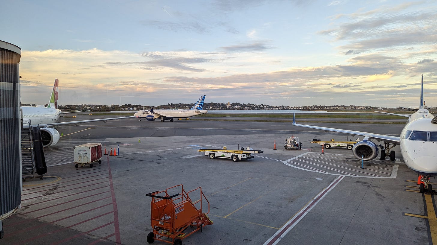 A photo out the window of an airport. There are two parked jets and one taxiing in the background. The sun is beginning to set.