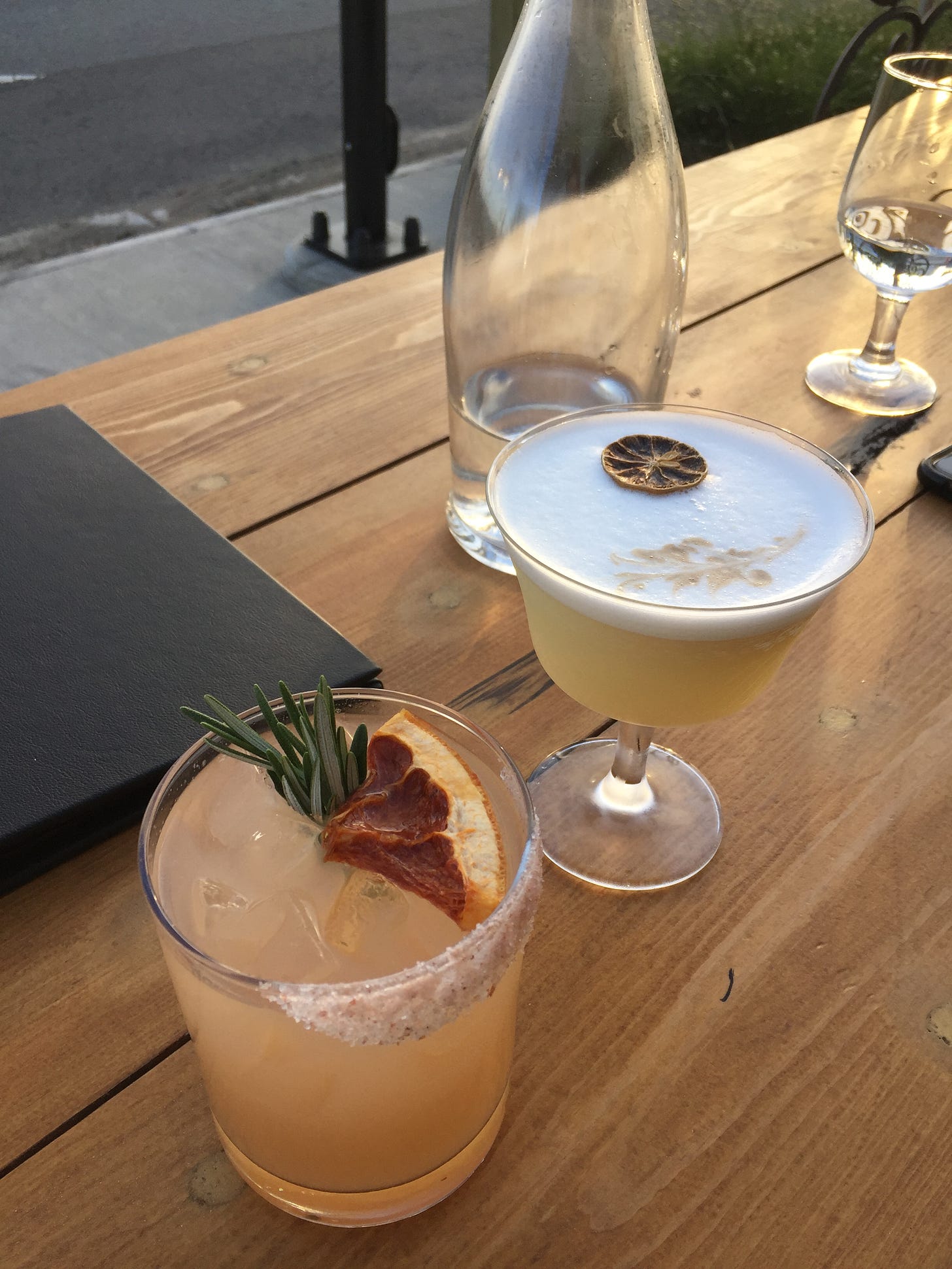 On a wooden outdoor bar are two cocktails in the foreground amongst a black menu, a large glass bottle of water, and water glasses. In a short glass is a light pink Paloma with a sprig of rosemary and dehydrated grapefruit, and a salted rim; and in a coupé glass is a foamy sour with a dried lime slice and a leaf drawn in the foam with bitters.