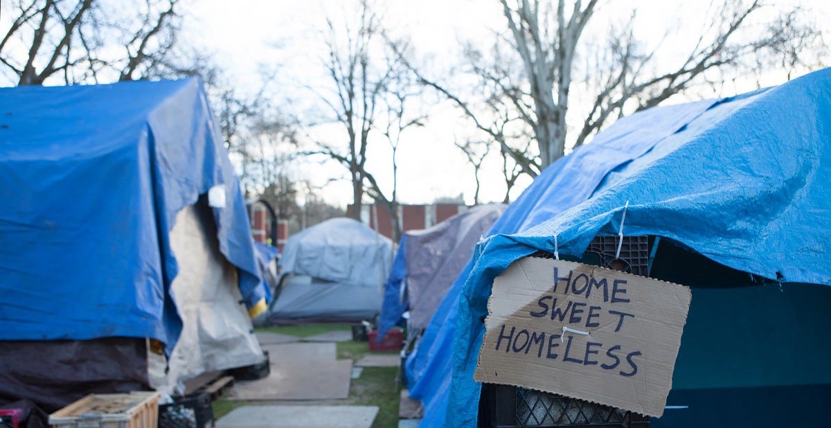 Image of a tent city with blue tarp covered tents. A tent in the foreground has a cardboard sign hanging from it