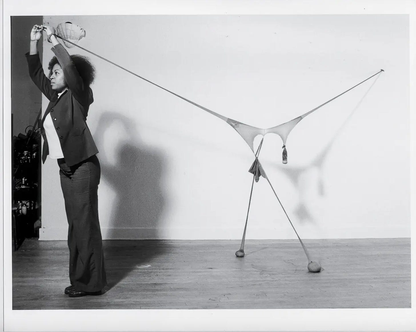 Black and white photo of nylon sculpture with pouches of weighted material affixed to a white wall on one end and suspended by Senga Nengudi at the other end. A shadow of the installation-performance is casted onto the wall. 