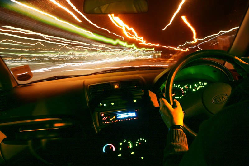 Image description: This is a color photo that depicts a driver in a car at night with their hand on the steering wheel as traffic lights glimmer in the distance. 